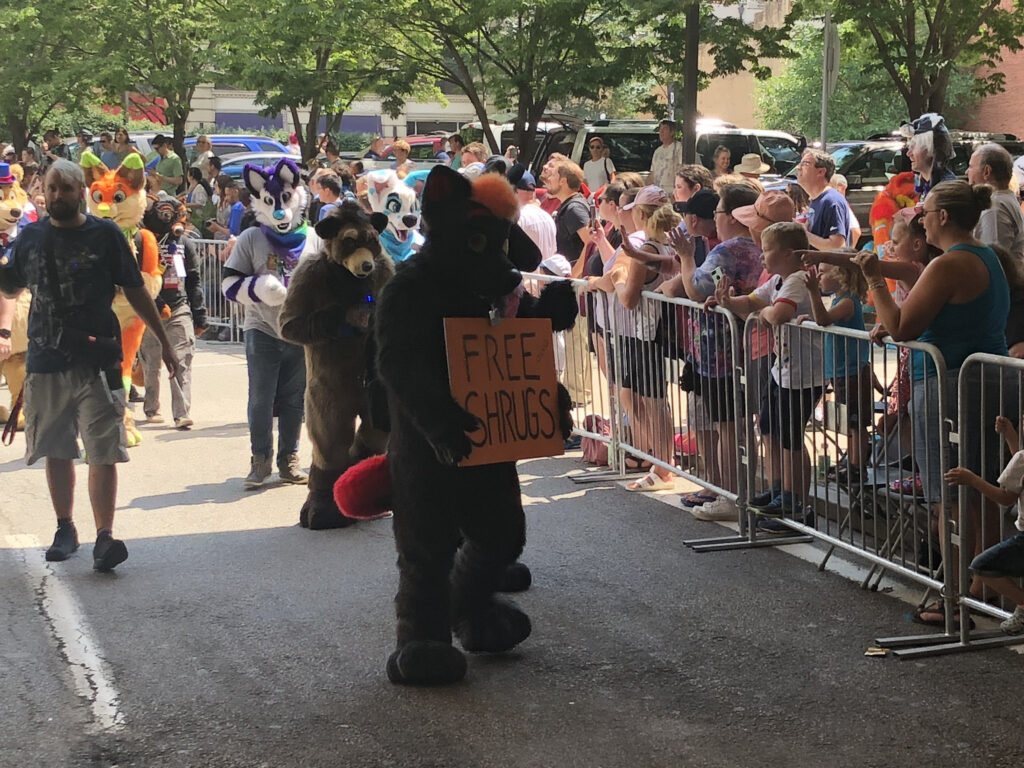 Fursuiter sfilano in strada durante una parata mentre il pubblico li osserva dietro le transenne; uno regge un cartello con scritto Free Shrugs