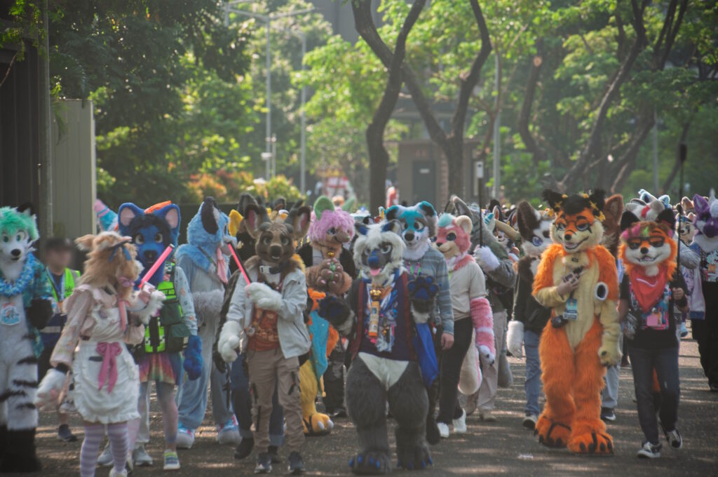Un lungo corteo di persone in costumi da animali antropomorfi colorati cammina all'aperto lungo un viale alberato